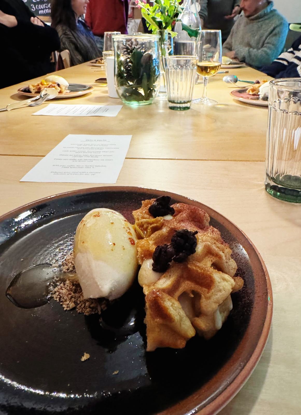 a dessert on a dark plate at a long shared table, with guests, menus and glasses behind it