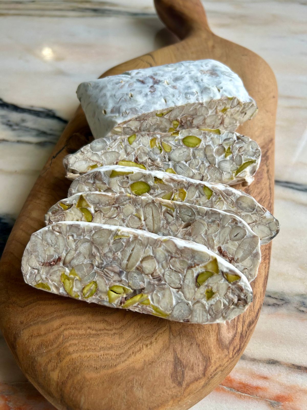 a log of patu tempeh cut into slices on an olive wood board, showing beans and pistachio inside